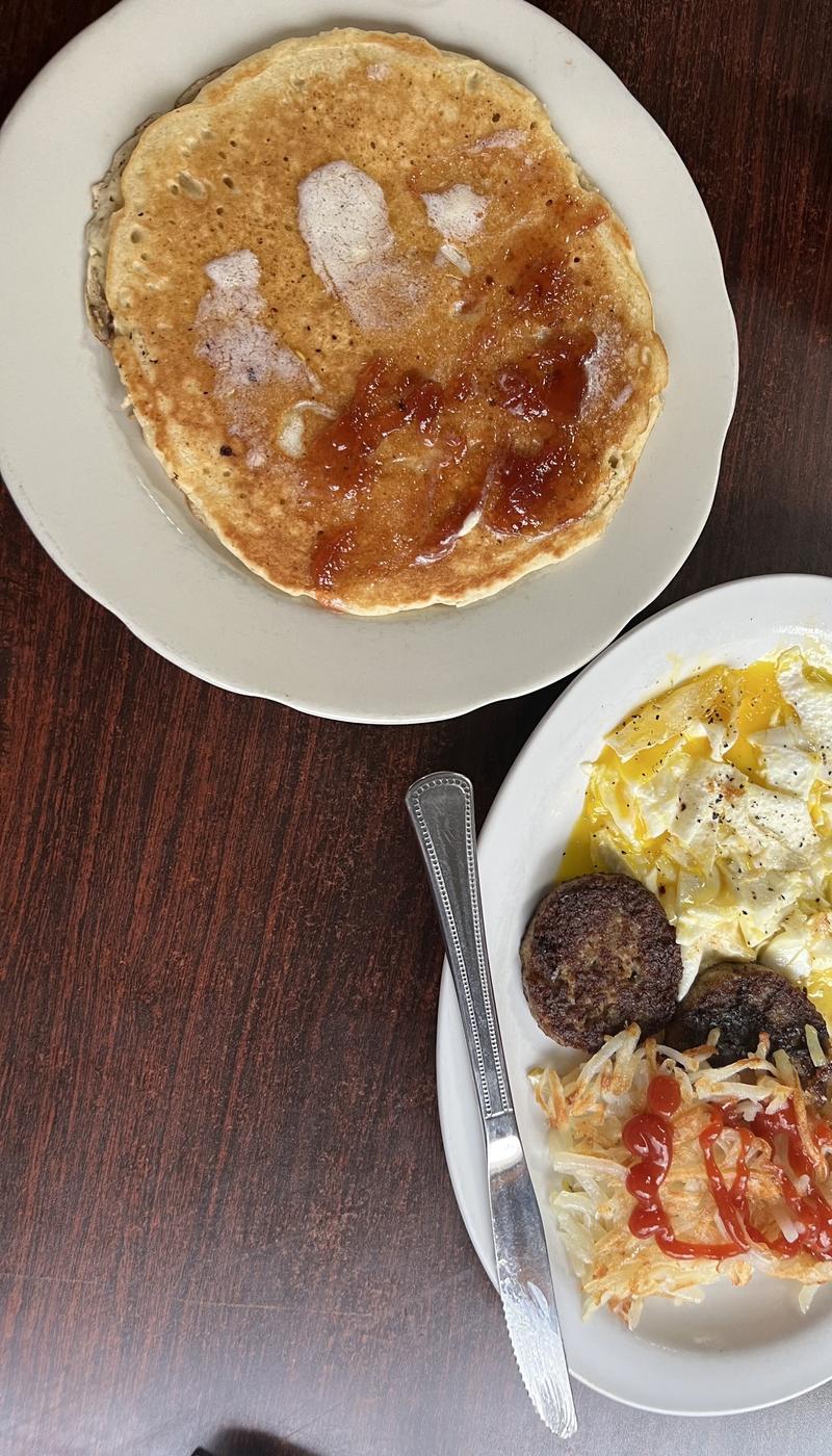 Breakfast on two plates: a pancake the width of the plate with butter and jelly, alongside over-easy eggs, two sausage patties and hash browns with ketchup.