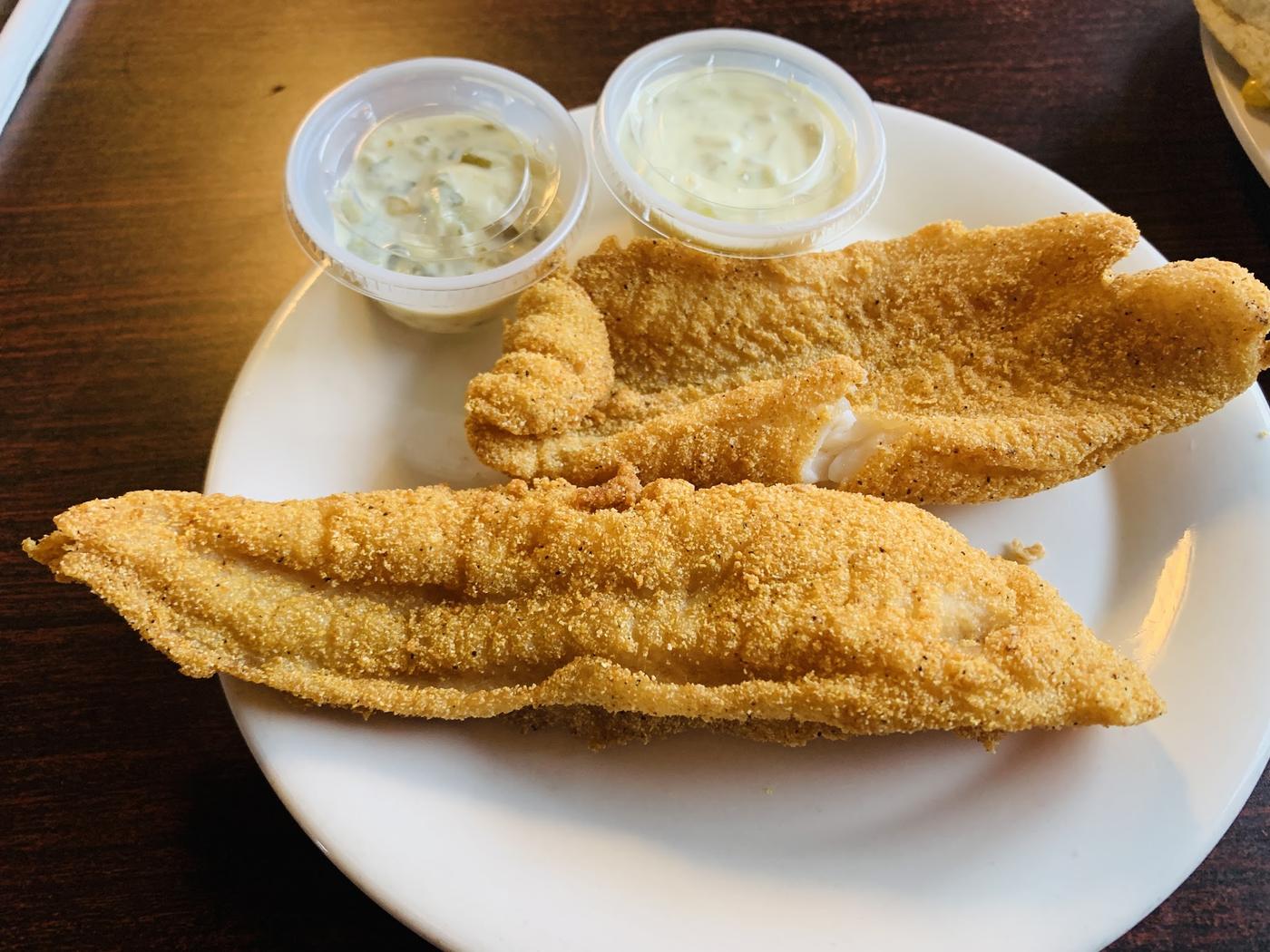 Two long cornmeal-breaded white fish fillets on a plate with two cups of tartar sauce.