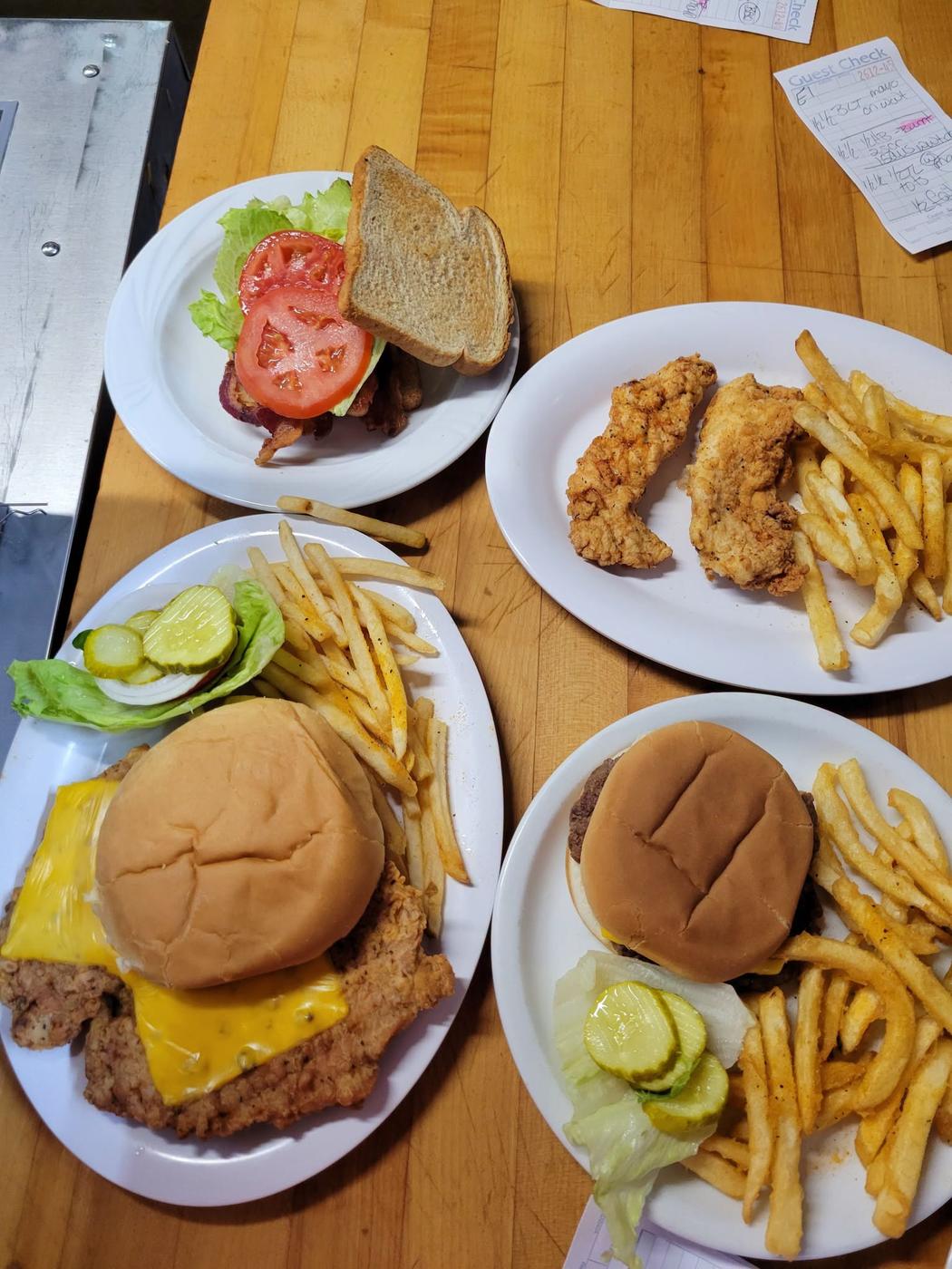 Four plates waiting on the kitchen pass: a BLT on toast, hand-breaded chicken strips with fries, a cheeseburger, and a breaded tenderloin topped with melted cheese.